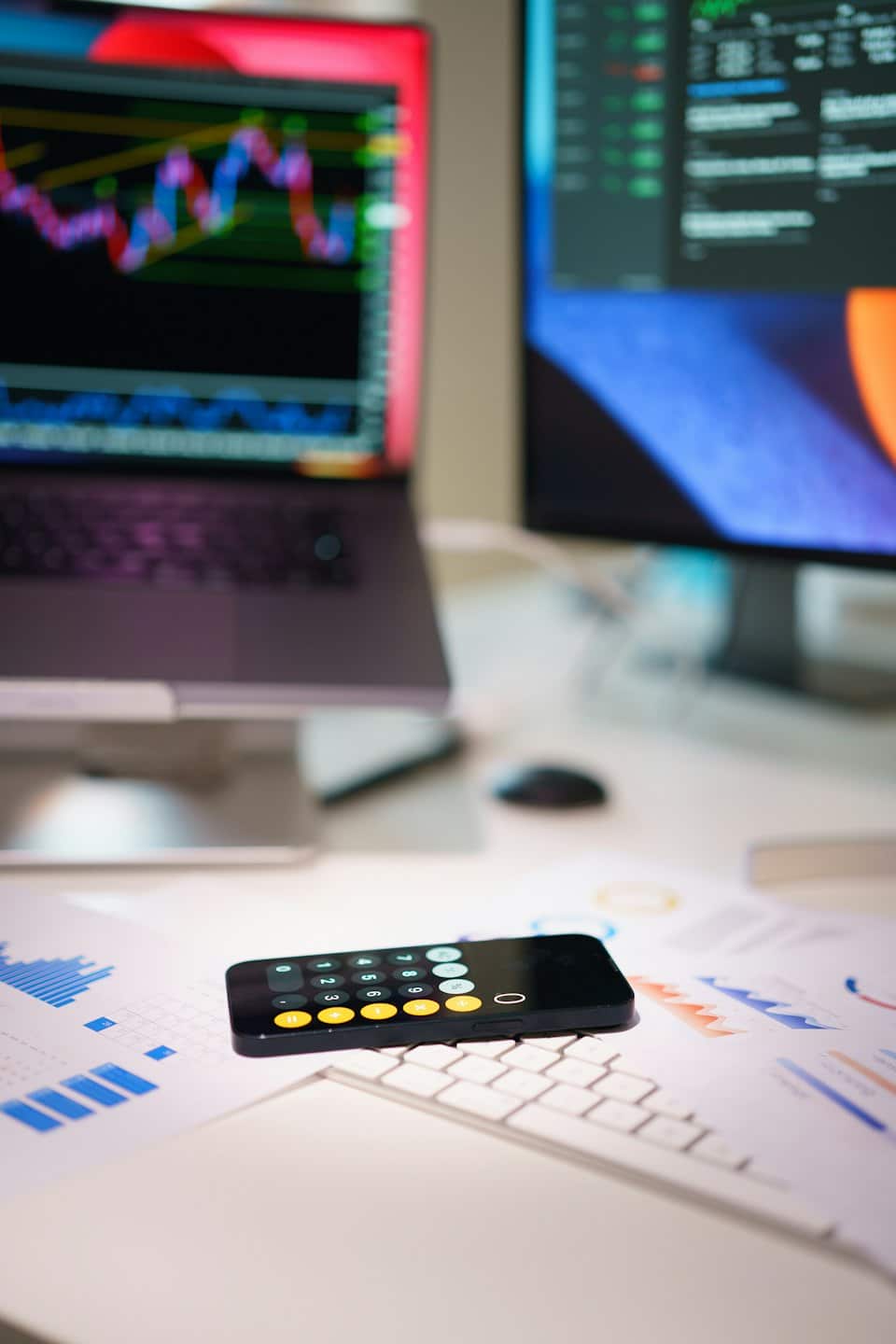 Smartfon na dokumentach finansowych obok laptopa z wykresami giełdowymi.