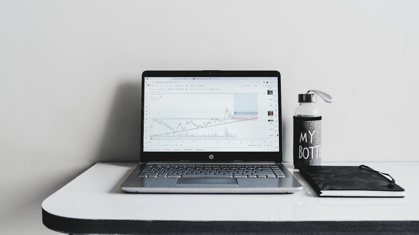 Na biurku leży laptop z wykresami finansowymi, obok niego zeszyt i butelka z wodą.